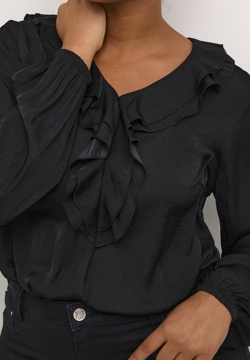 Blouse noire à volants avec manches longues bouffantes portée par une personne avec la main près du cou et de la taille, sur un fond uni.