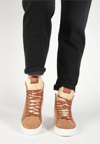 Ruskeat mokkakengät korkealla varrella, joissa on valkoiset kumipohjat, vaaleanruskea turkkivuori ja ruskeat nauhat; mustat farkut, jotka on käännetty nilkasta, parantavat näkyvyyttä.