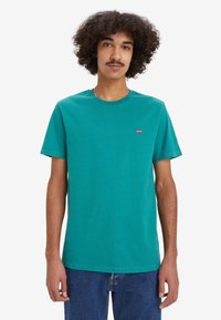 Teal bomba majica iz bombaža s krojem okroglega vratu in kratkimi rokavi, na levi strani z majhnim rdečim logotipom "Levi's". Preprost dizajn, brez vzorcev.