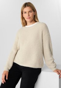 Jasnobeżowy sweter dzianinowy o ribbowanej fakturze, okrągłym dekolcie i długich rękawach. Zestawiony z czarnymi spodniami, ma swobodny i casualowy charakter.