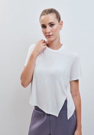 Jeune femme aux cheveux tirés en arrière, portant un haut blanc manches courtes à ourlet asymétrique et un pantalon gris, debout devant un fond uni.