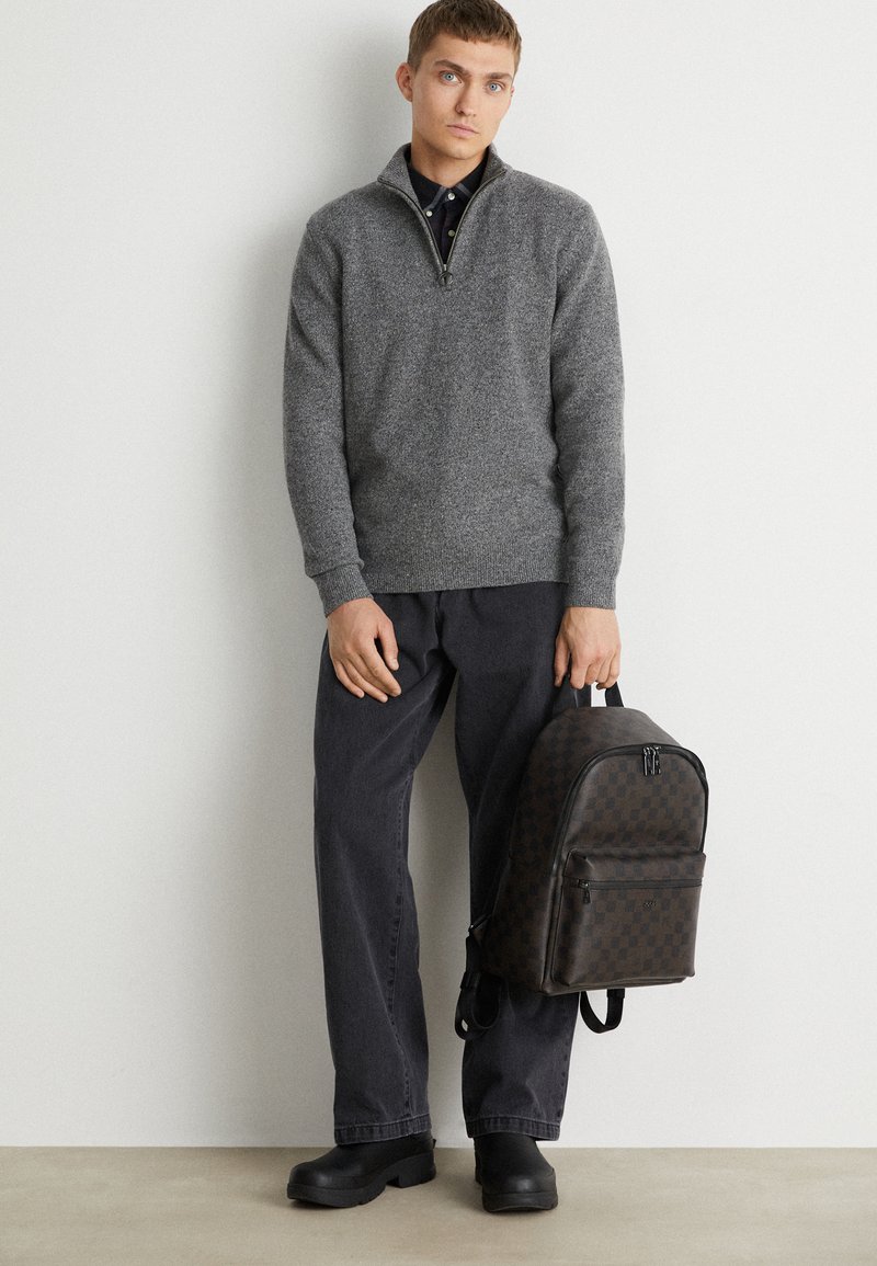 Grauer Strickpullover mit halbem Reißverschluss am Hals, dunkelgraue Hose und ein brauner Rucksack im Schachbrettmuster mit einer Vordertasche und zwei Griffen.