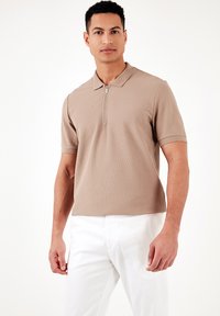 Bežs polo krekls ar rāvējslēdzēja kakla izgriezumu un īsām piedurknēm, ar strukturētu adījumu un brīvu piegriezumu, kombinēts ar baltām biksēm.