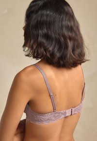 Nėriniuota liemenėlė šviesiai mauvės spalvos su banguotu kraštu, reguliuojamomis petnešėlėmis ir užsegimu gale, puošta dekoratyviniais aparatūros akcentais.