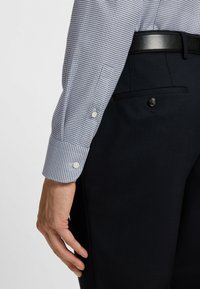 Camicia a quadri blu e bianca con polsini a bottone, abbinata a pantaloni neri testurizzati con tasca posteriore e dettaglio a bottone.