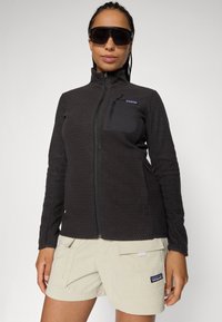 Vrouw in een zwart, textuur ritsjack met borstzak en beige shorts, accessorized met grote zwarte zonnebril op een effen achtergrond.
