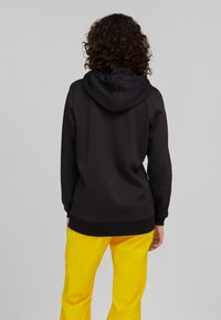 Sudadera negra con capucha y bolsillo canguro, combinada con pantalones amarillos brillantes. La prenda presenta una textura suave y un ajuste relajado.