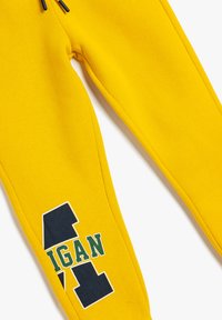 Gule sweatsbukser med et navy og grønt print med bogstavet "A" og ordet "MIGAN." Lavet af blødt bomuld, med en afslappet pasform og snøre.