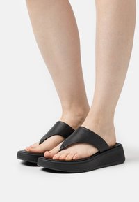 Sandali flip-flop neri in pelle con una fascia ampia e angolata e una suola piatta nera spessa. Le dita dei piedi sono visibili e mostrano un colore naturale delle unghie.