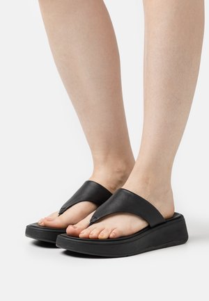 Des sandales en cuir noir, type tongs, avec une large bride angulaire et une semelle épaisse noire. Les orteils exposés révèlent une couleur de vernis naturel.