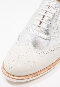 Chaussure brogue classique en cuir blanc et argent avec perforations décoratives et semelle cousue marron clair.