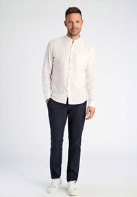 Camicia chiara a maniche lunghe con taschino, abbinata a pantaloni scuri e scarpe da ginnastica bianche, in piedi su uno sfondo neutro.