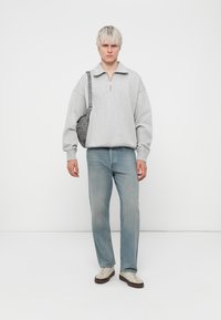 Harmaa fleece-huppari, jossa on puolivetoketju ja löysä malli. Vaaleansiniset suorat farkut ja kermaiset lenkkarit ruskeilla pohjilla. Harmaa olkalaukku.