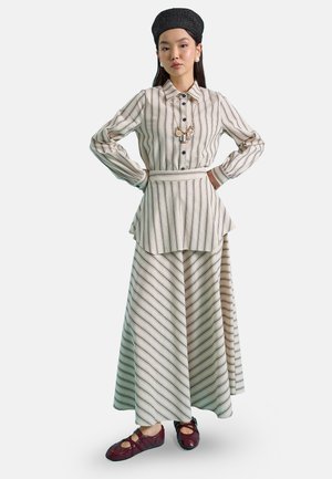Femme debout les mains sur les hanches, portant une chemise à manches longues rayée, un pantalon large assorti, un foulard noir sur la tête et des chaussures bordeaux.