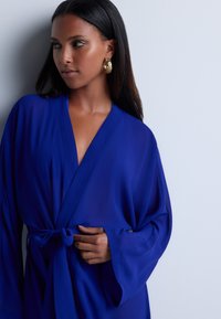 Robe bleu en tissu léger avec une taille à nouer et des manches larges, dotée d'un décolleté en V profond et d'une texture lisse.