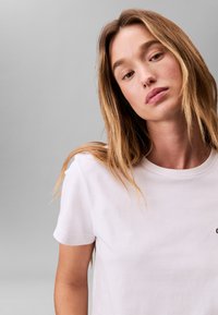 Weißes Baumwoll-T-Shirt mit rundem Ausschnitt und kurzen Ärmeln, mit einem kleinen schwarzen Logo auf der linken Brustseite. Glatte Textur.