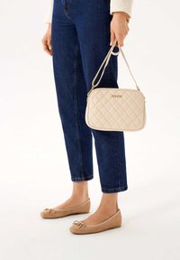 Borsa a tracolla beige trapuntata con chiusura a zip e logo dorato, tenuta da una persona che indossa jeans blu scuro e ballerine beige.