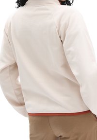 Chaqueta de forro polar de color beige claro con cuello alto y mangas raglán. Presenta un dobladillo de color óxido en la cintura.