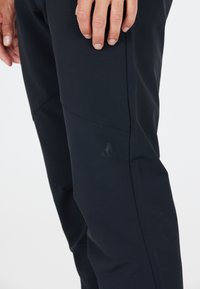 Svarta soft-shell byxor har en slim fit, en artikulerad knädessign och en diskret logga på låret, med en mjuk, något glänsande textur.