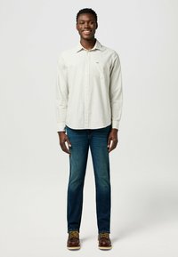 Camicia bianca a maniche lunghe con strisce verdi, abbottonata, con taschino sul petto. Indossata con jeans blu scuro e stivali marroni.