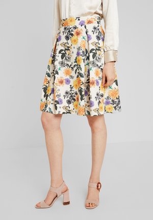 Vrouw draagt een geplooide witte rok met oranje, paarse en zwarte bloemmotieven, gecombineerd met beige sandalen met hakken en een crèmekleurige blouse.