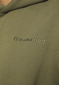 Close-up of olive green fabric with the word "Humanrace" embroidered in matching thread on the chest area of clothing.