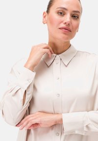 Blusa beige chiaro con colletto classico, chiusura a bottoni, maniche lunghe con polsini con bottoni e dettagli laterali discreti.