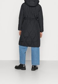 Černý prošívaný kabát s kapucí, stažený v pase a se stehovaným vzorem. Kombinován se světle modrými širokými džíny a černými robustními botami.