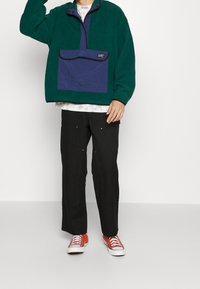 Grön fleecepullov med en marinblå framficka och en halvdragkedjekrage, i kombination med svarta byxor och röda sneakers. Strukturerad, avslappnad design.