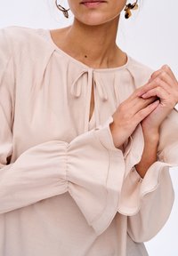 Jasnoróżowa bluzka z okrągłym dekoltem z ozdobą w postaci wiązania. Rękawy są bufiaste i rozszerzane, o miękkiej teksturze i subtelnym połysku.