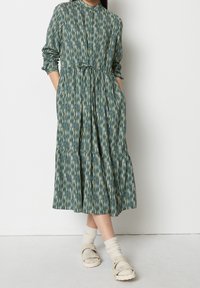 Vestito verde con motivo a righe verticali, chiusura a bottoni frontale, maniche lunghe, vita cinte e gonna a strati. Indossato con calzini leggeri e sandali.