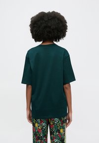 Mørkegrøn bomuld T-shirt med korte ærmer, afslappet pasform og rund halsudskæring, vist bagfra. Kombineret med mønstrede blomstrede bukser.