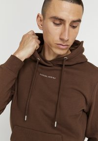 Brun hættejakke med en frontlomme, snøre med sølvspidser og hvid tekst, der står "CASUAL FRIDAY" på brystet. Glat tekstur.