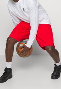 Rote Sportshorts mit einem schwarzen Nike-Logo, kombiniert mit einem weißen Langarmshirt. Die Person hält einen mehrfarbigen Basketball.