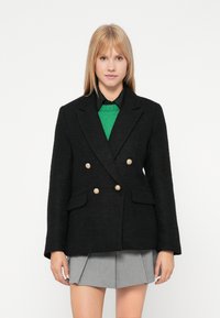 Črn dvoredni srajčni blazer z zlatimi gumbi, teksturirana tkanina in dva sprednja žepa; kombiniran z zelenim puloverjem in pregibno sivo krilo.