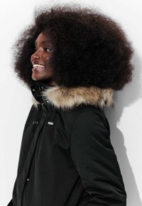 Parka noire avec une capuche bordée de fourrure, dotée d'une fermeture éclair et de boutons-pression, ainsi que de cordons de serrage au niveau du cou. Texture lisse avec une finition mate.