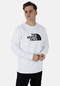 Sudadera blanca de algodón, con puños y dobladillo de canalé, que presenta un diseño de logo negro de "The North Face" en el frente.