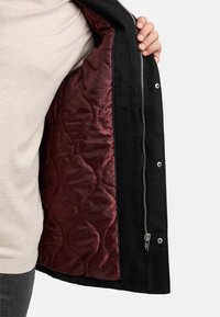 Svart jakke med quiltet burgunderfôr med prikkete mønster. Glidelås og trykknapper synlige foran.