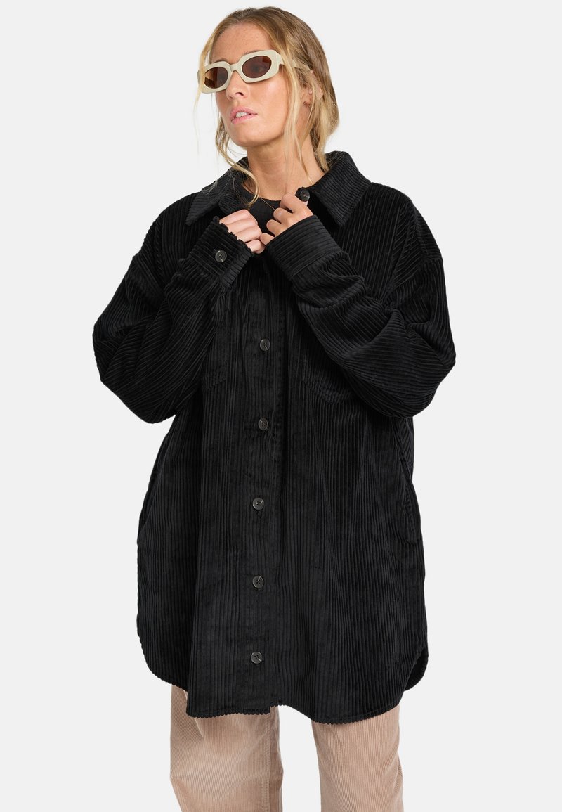 Schwarze Cordjacke mit großem Kragen, frontalen Knöpfen und zwei seitlichen Taschen; weist durchgehend strukturierte vertikale Rippungen auf.