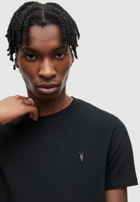 Černé bavlněné tričko s kulatým výstřihem a krátkými rukávy. Má malou šedou lebku kozy vyšitou na levé hrudi. Hladká textura.