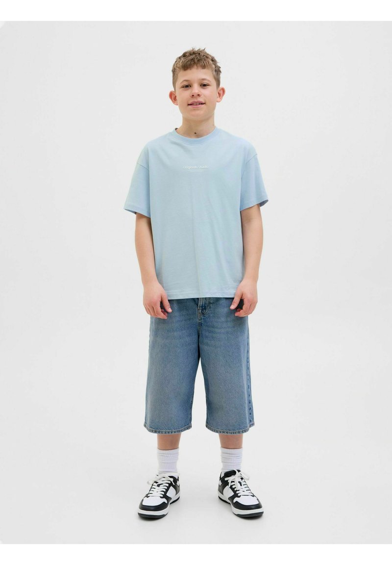 Světle modré tričko s krátkým rukávem a logem na hrudi, v kombinaci s širokými modrými džínovými šortkami, bílými ponožkami a černo-bílými teniskami.