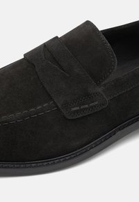 Mocassin en daim noir avec un design cousu. Comprend une bride décorative et une texture lisse, avec une semelle en caoutchouc pour l'adhérence.