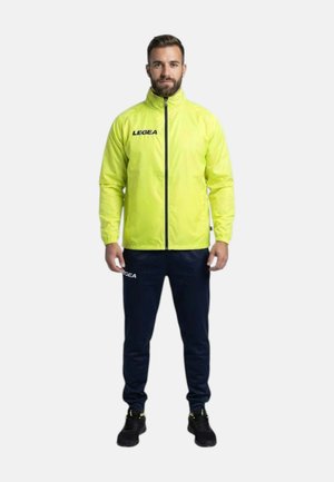 Uomo in piedi che indossa una giacca gialla brillante con zip e pantaloni blu scuro con sneakers nere su uno sfondo bianco uniforme.