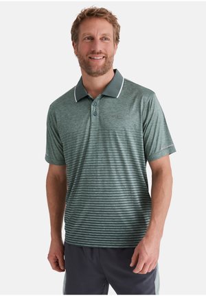 Mann trägt ein grünes, gestreiftes Poloshirt mit Kragen, dunkle Shorts, lächelt und schaut leicht nach links vor einem einfarbigen Hintergrund.