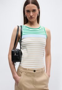 Νεαρή γυναίκα που φοράει ένα αμάνικο ριγέ τοπ με πράσινες, μπλε και μπεζ γραμμές, μπεζ παντελόνι με τα χέρια στις τσέπες και μια μαύρη τσάντα ώμου.