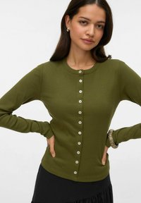Mlada žena koja nosi maslinasto zelenu bluzu s dugim rukavima i gumbima, srebrne naušnice te prstenastu narukvicu s prugama na zapešću.
