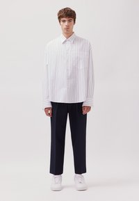 Camicetta bianca con righe verticali blu, maniche lunghe e taschino sul petto, abbinata a pantaloni navy scuri e scarpe da ginnastica bianche.