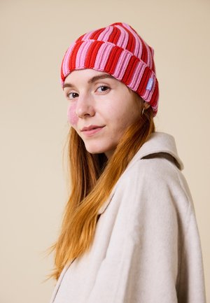 Knit beanie in red and pink stripes, featuring a folded hem and logo tag. Soft texture, fitted design. Model wears a beige coat.