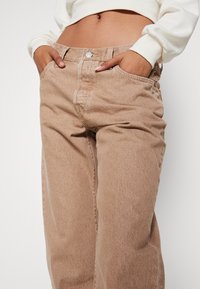 Personne portant un jean taille haute beige les mains dans les poches et un sweat-shirt blanc court, montrant un piercing au nombril.