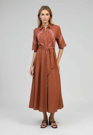 Femme portant une robe midi couleur rouille avec broderies de feuilles roses, devant boutonné, taille ceinturée, et sandales à talons sur un fond neutre.